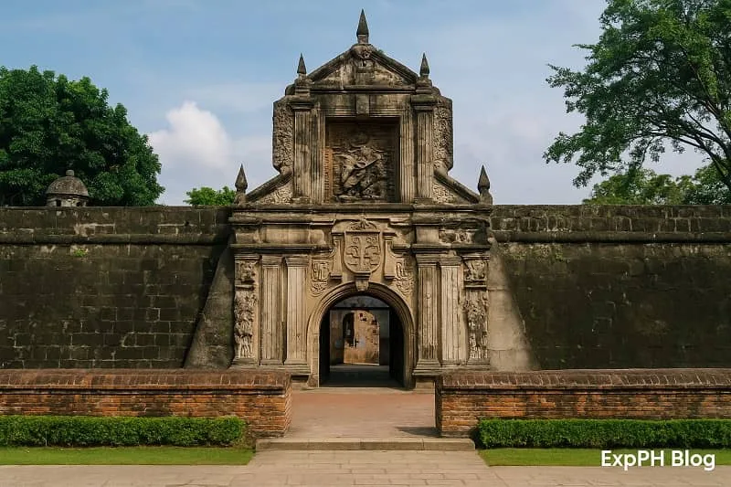Realistic view of Fort Santiago’s stone gate in Manila with detailed carvings, surrounding walls, green trees, clear sky, and the ExpPH Blog logo at the bottom corner.