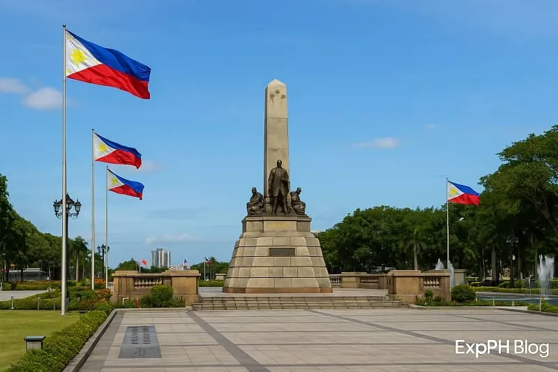 Realistic view of the Rizal Monument at Rizal Park in Manila with Philippine flags, green landscape, clear blue sky, and the ExpPH Blog logo at the bottom corner.